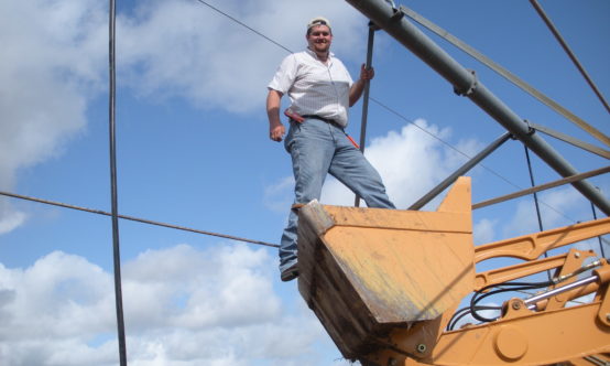 Charles Swanson holding onto a center pivot beam while balanced atop the raised claw of a tractor.